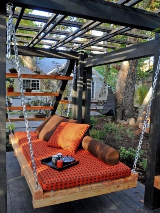 מיטה תלויה בחוץ בגינה בריפוד בצבע כתום מגש ועליו נר כוסות קפה